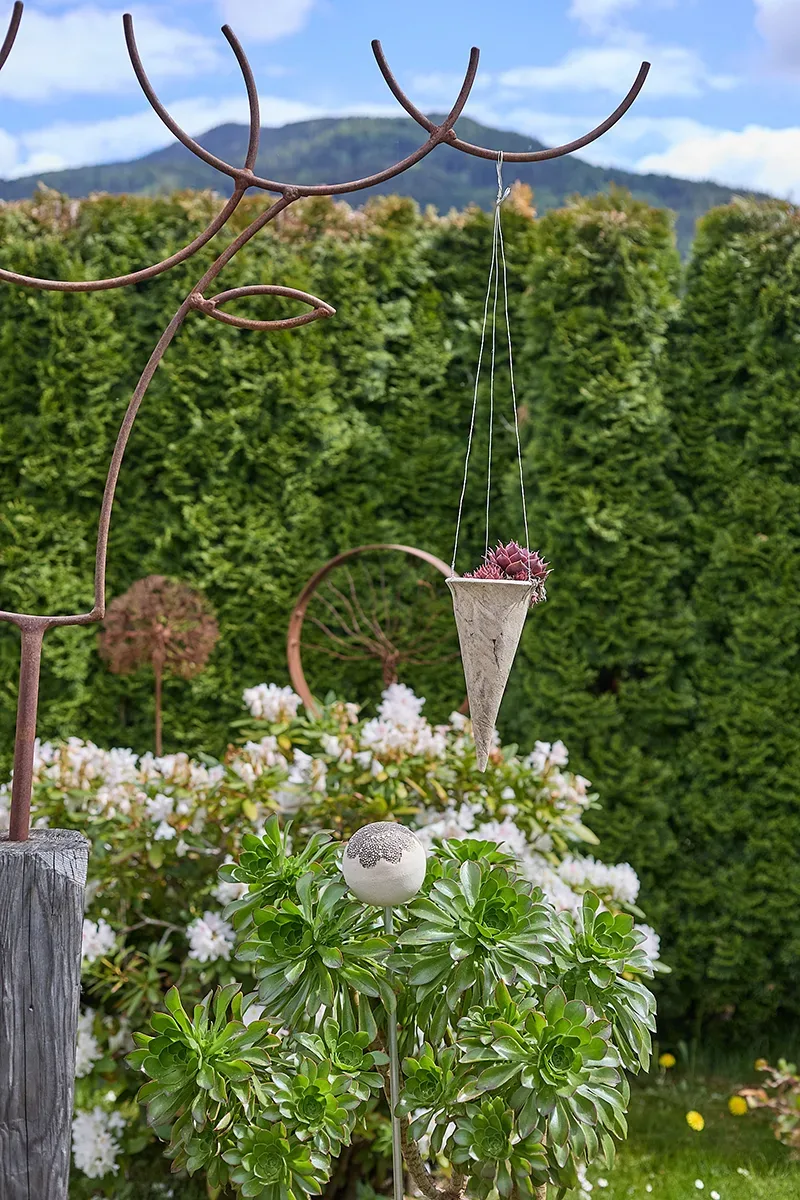 Metallskulptur mit hängendem Kegel, bepflanzt, im Garten vor Hecke und Blumen.