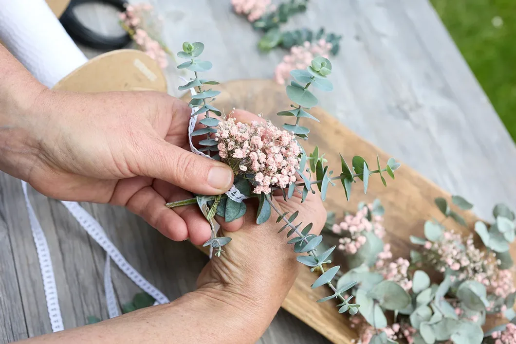 Hände binden Sträußchen aus Eukalyptus und rosa Blumen auf Holztisch.