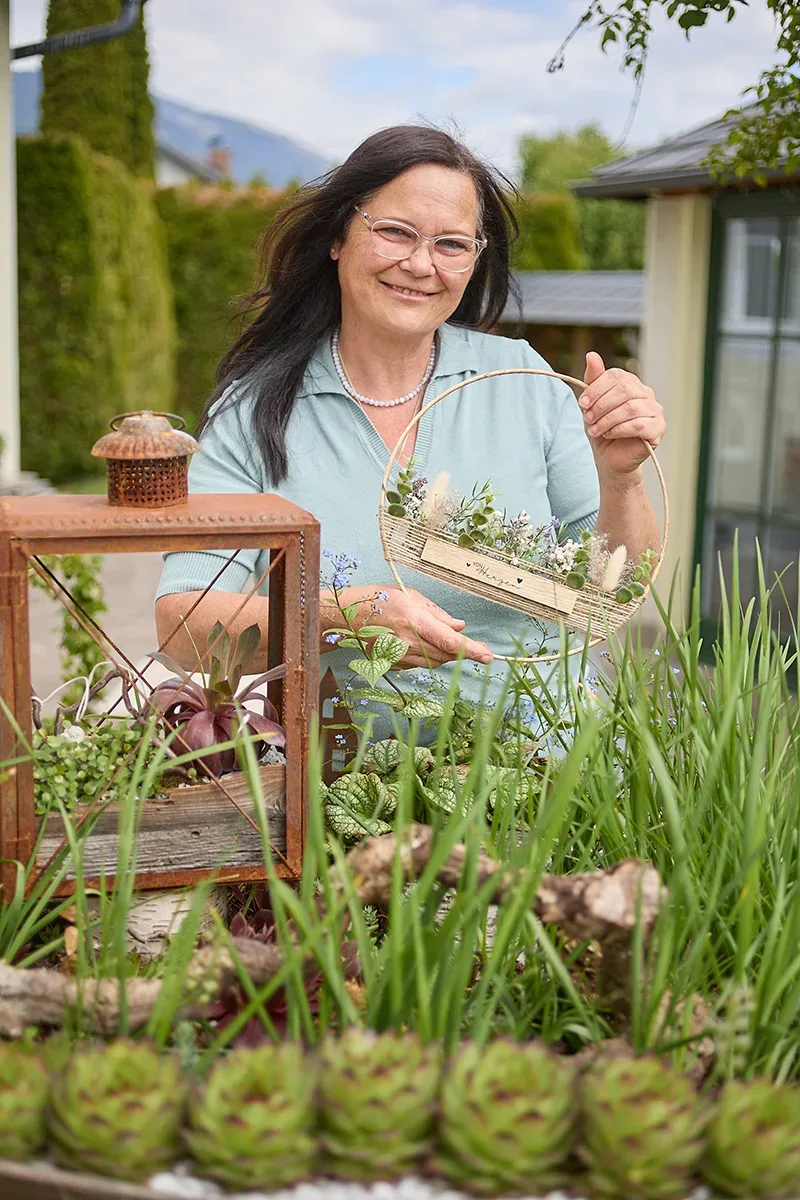 Frau in Garten hält florales Dekoelement neben Holzlaterne und Pflanzenarrangement.